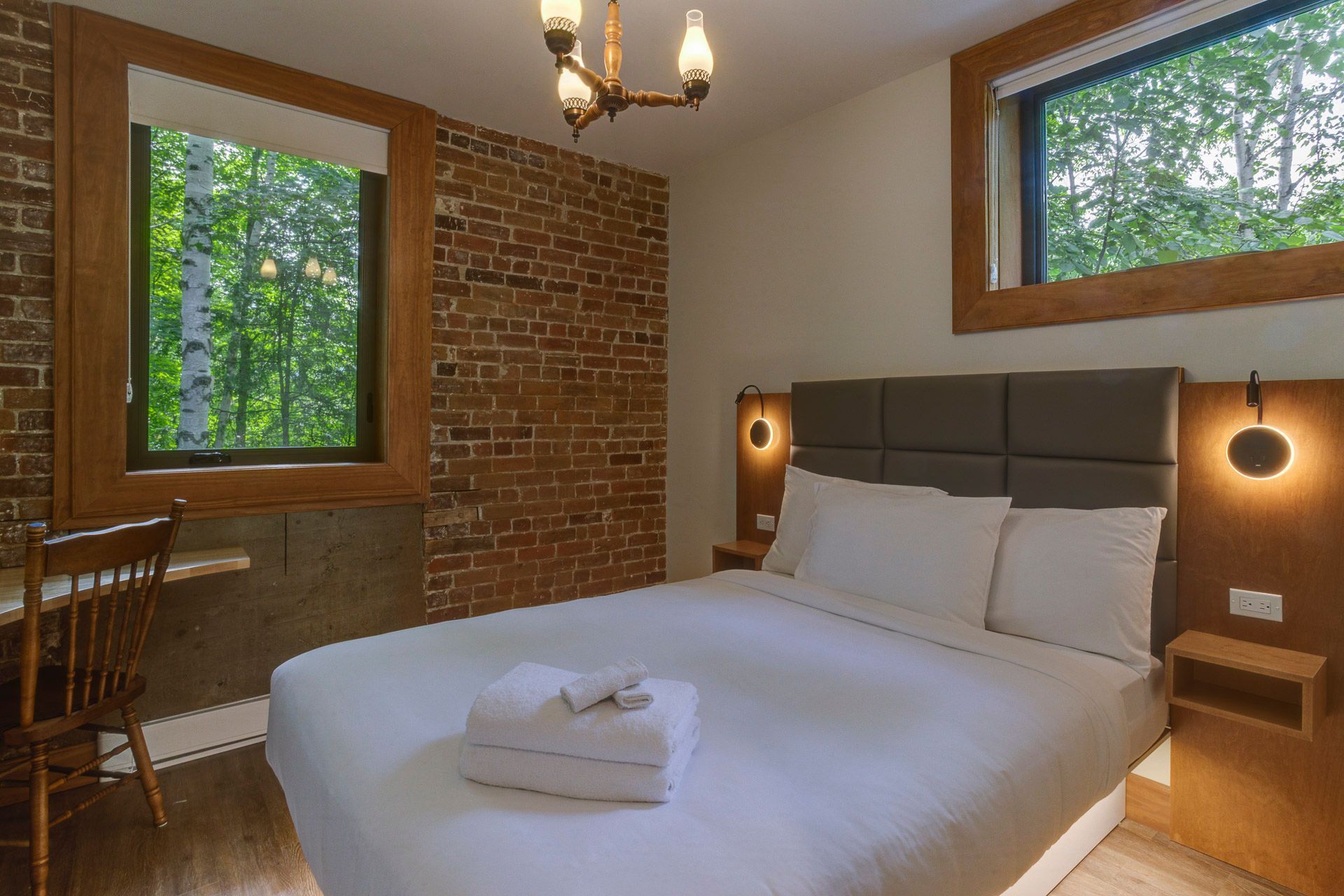 Chambre confortable avec un lit blanc, un mur de briques, des éléments en bois et deux fenêtres donnant sur les arbres