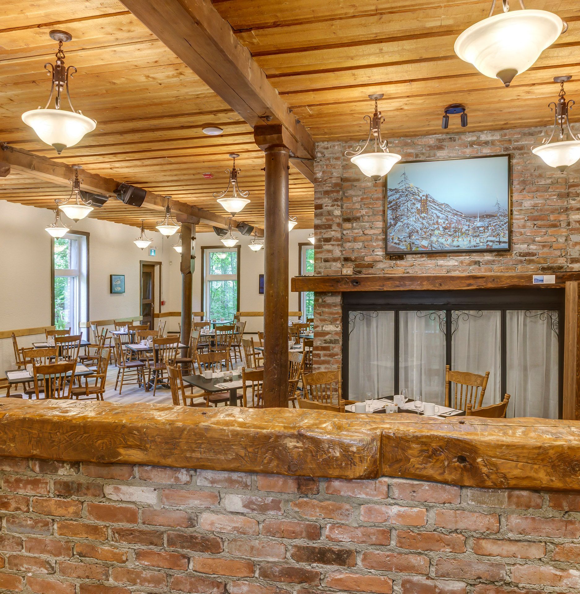 Salle à manger rustique avec poutres apparentes, murs en briques, lustres et tables disposées près d'une cheminée.