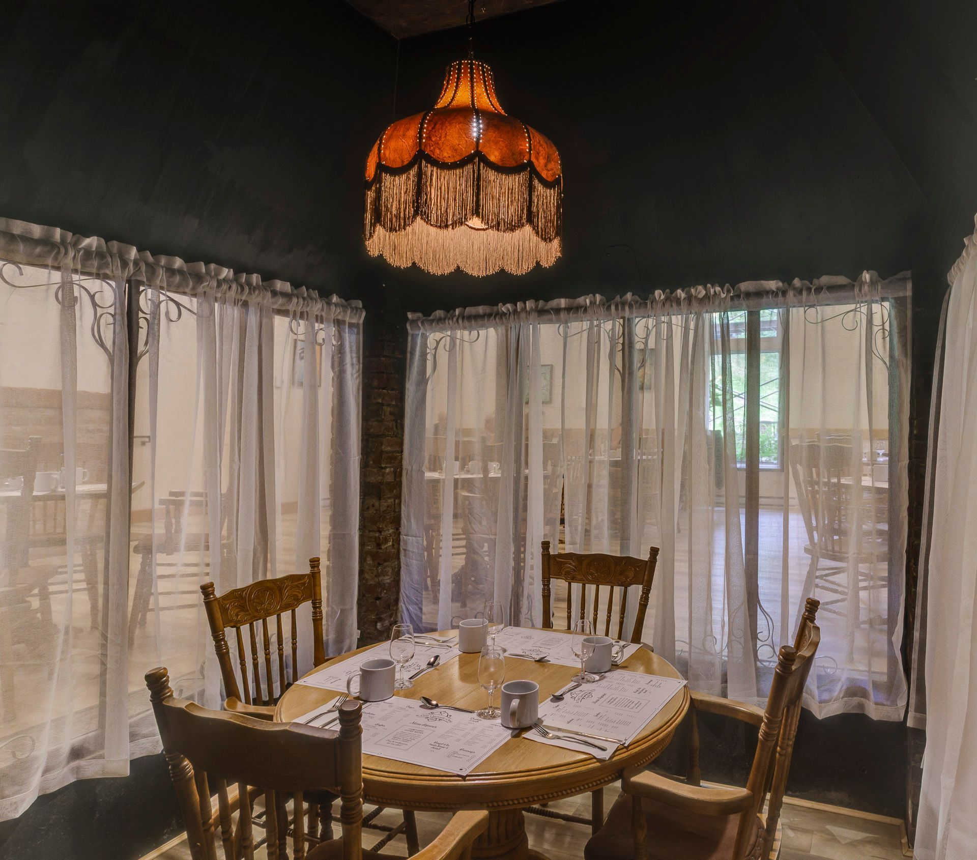 Salle à manger tamisée avec une table ronde, des chaises en bois, des rideaux en dentelle et une lampe suspendue.