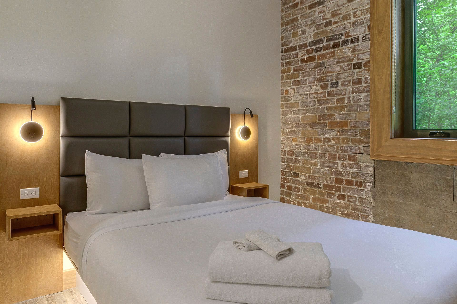 Chambre moderne avec un lit bien fait, des appliques murales, un mur en briques apparentes et une vue sur la verdure.