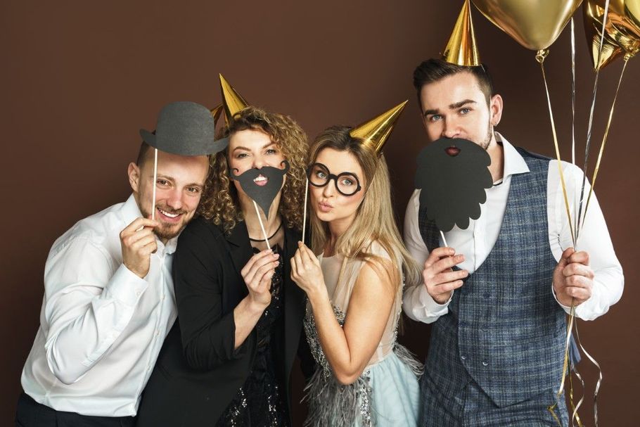 Four people in party hats pose with fake beards and glasses, holding gold streamers and smiling.