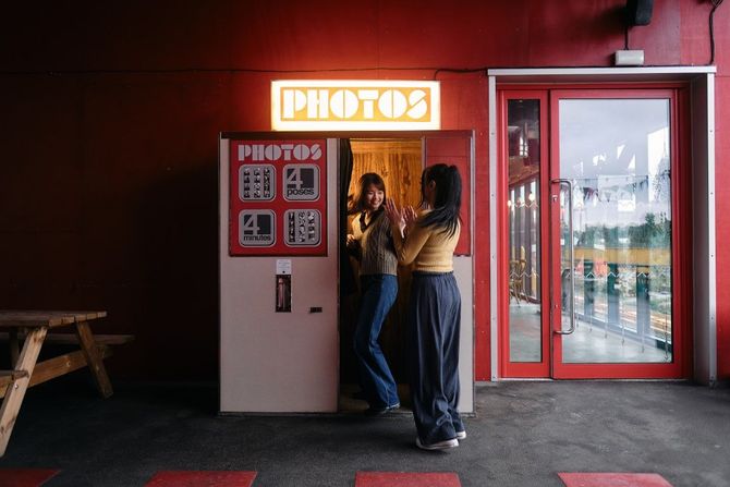 Two people posing in a red photo booth beside a lit “PHOTO” sign.