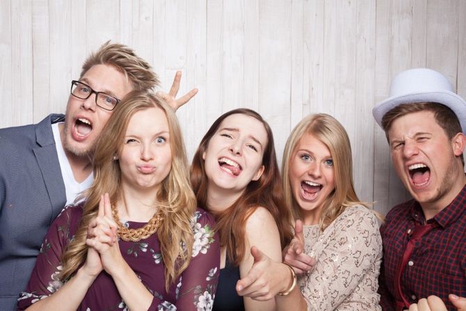 Five friends posing playfully with big smiles against a light wood wall