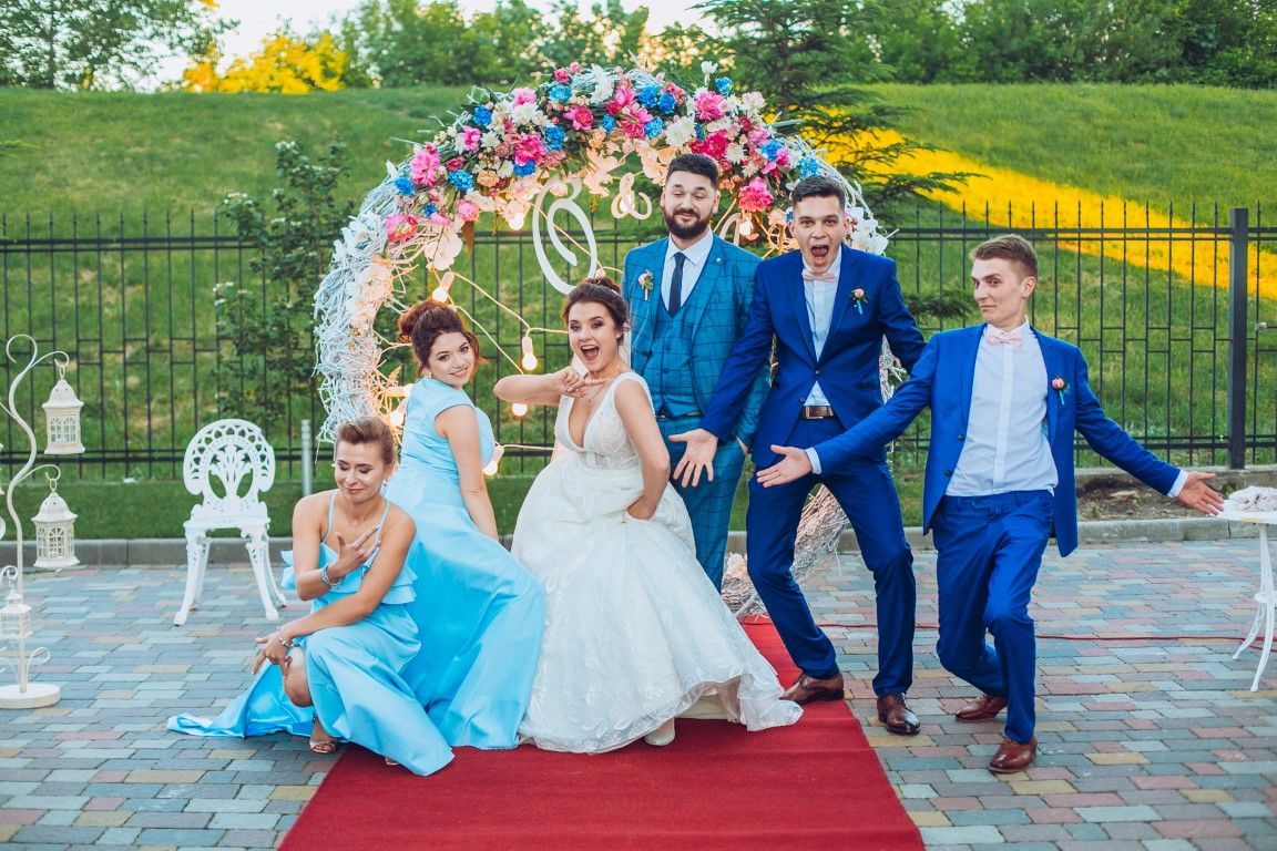 Wedding party posing on a red carpet outdoors beside a floral arch and fountain