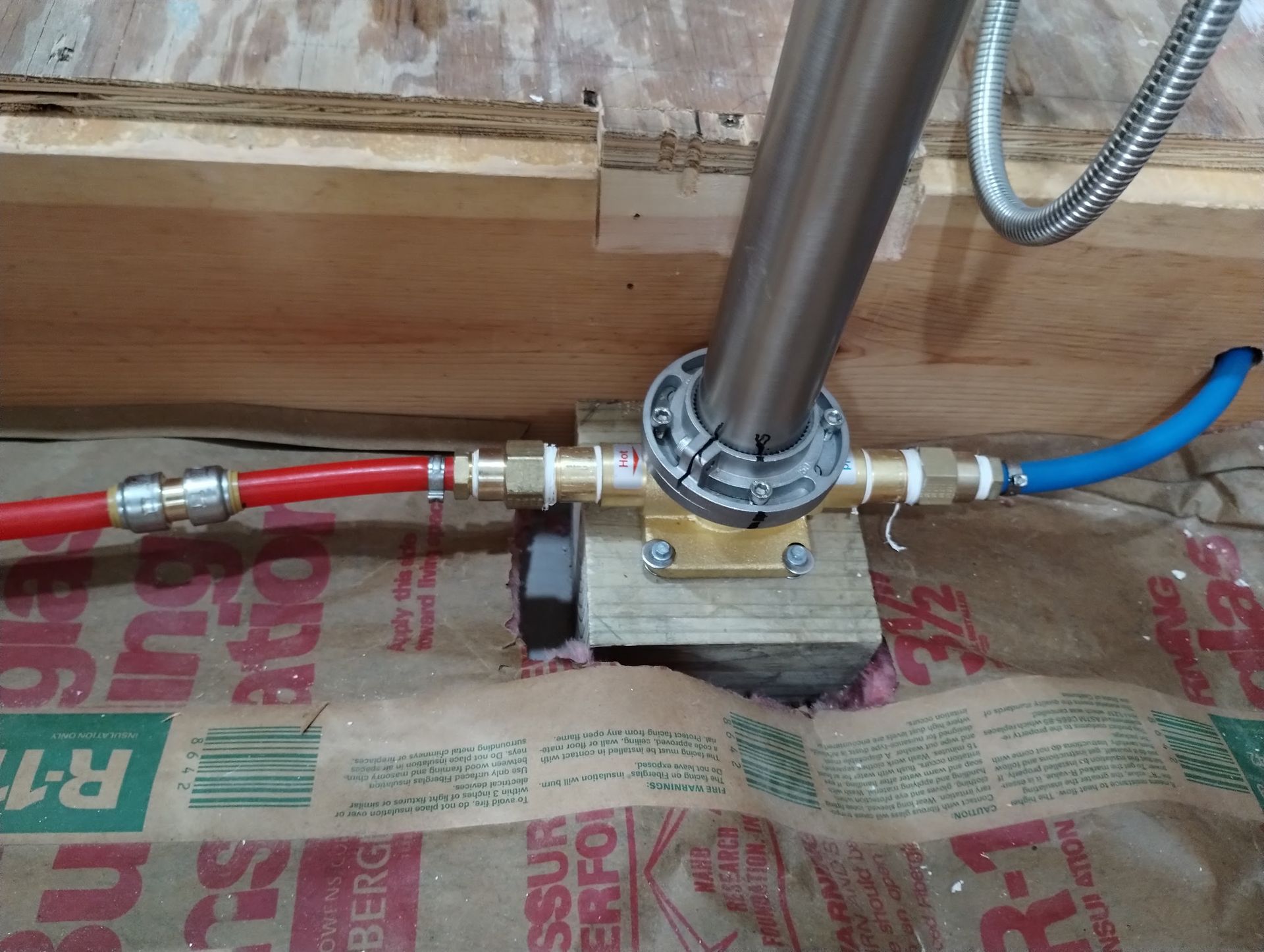 Close-up of metal plumbing pipe with red and blue hoses connected to a brass fitting on a wooden floor.