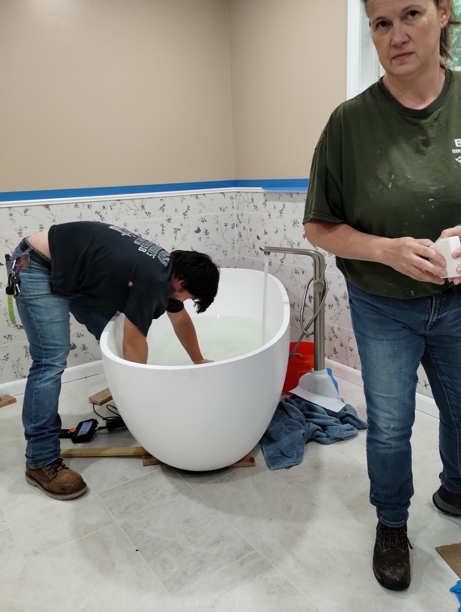 Two workers installing a large white bathtub in a tiled bathroom under construction