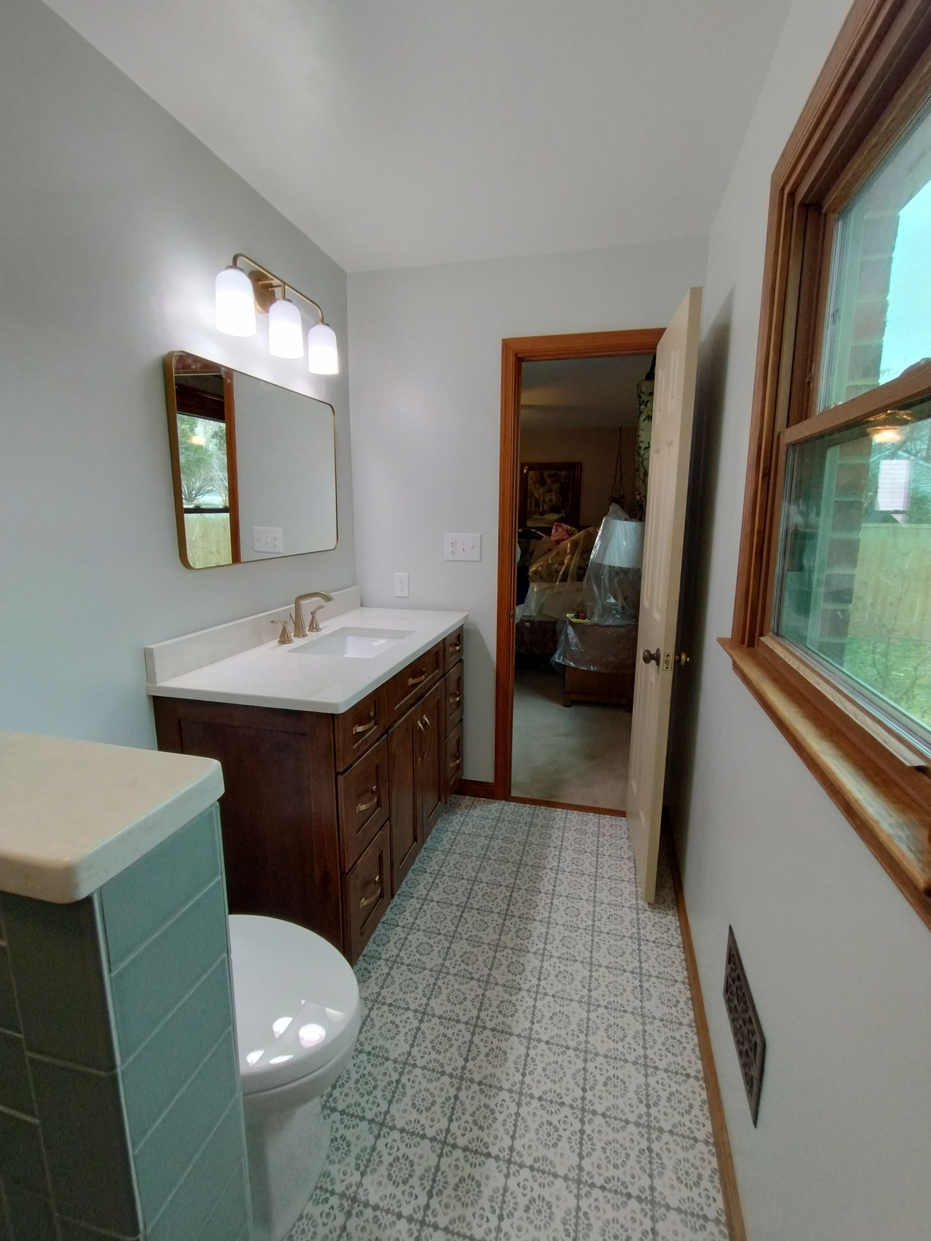 Narrow bathroom with a sink, mirror, toilet, patterned tile floor, and window with wood trim.