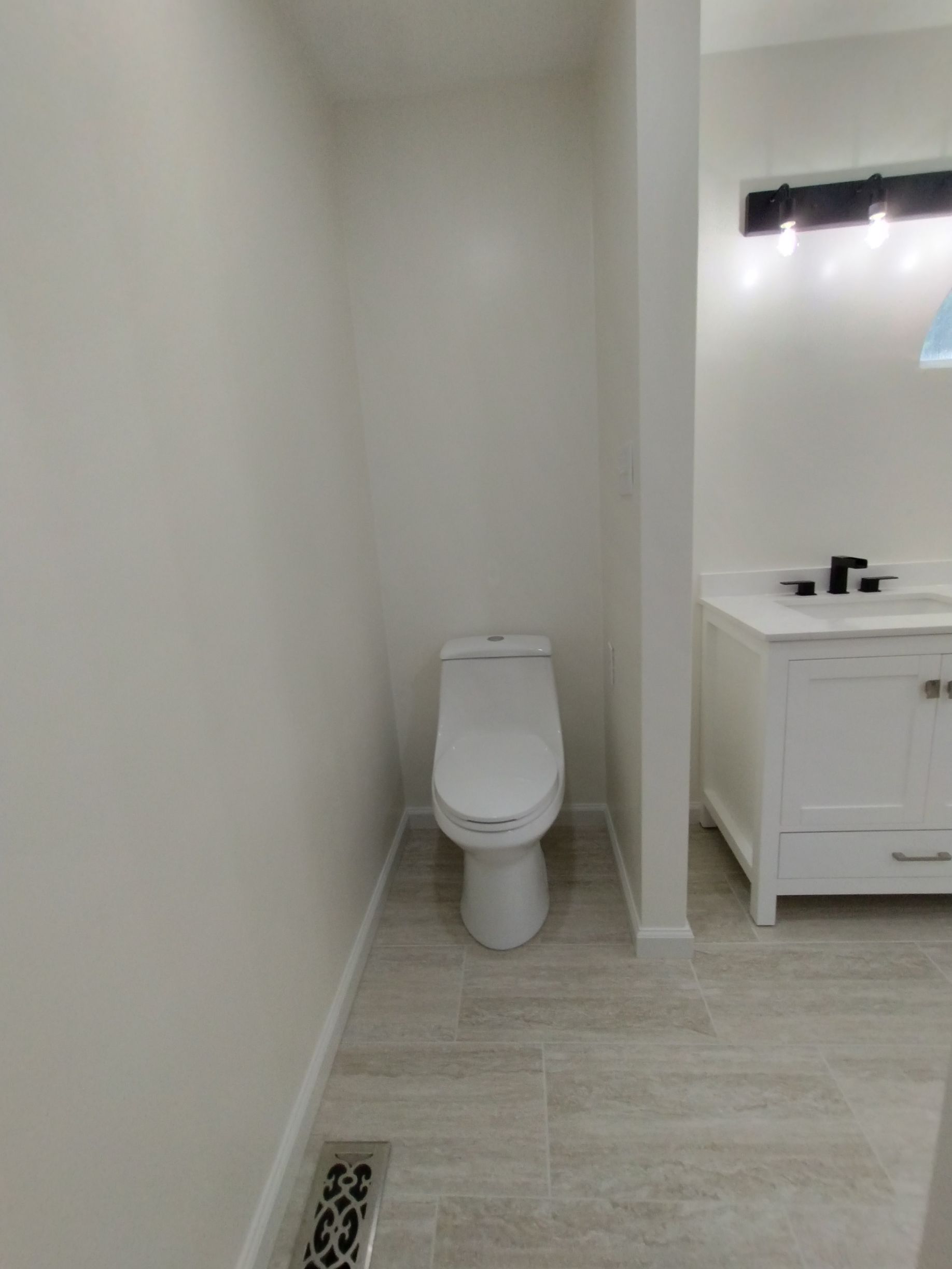 Small bathroom with a toilet beside a vanity and sink, light tile floor, and a floor vent.