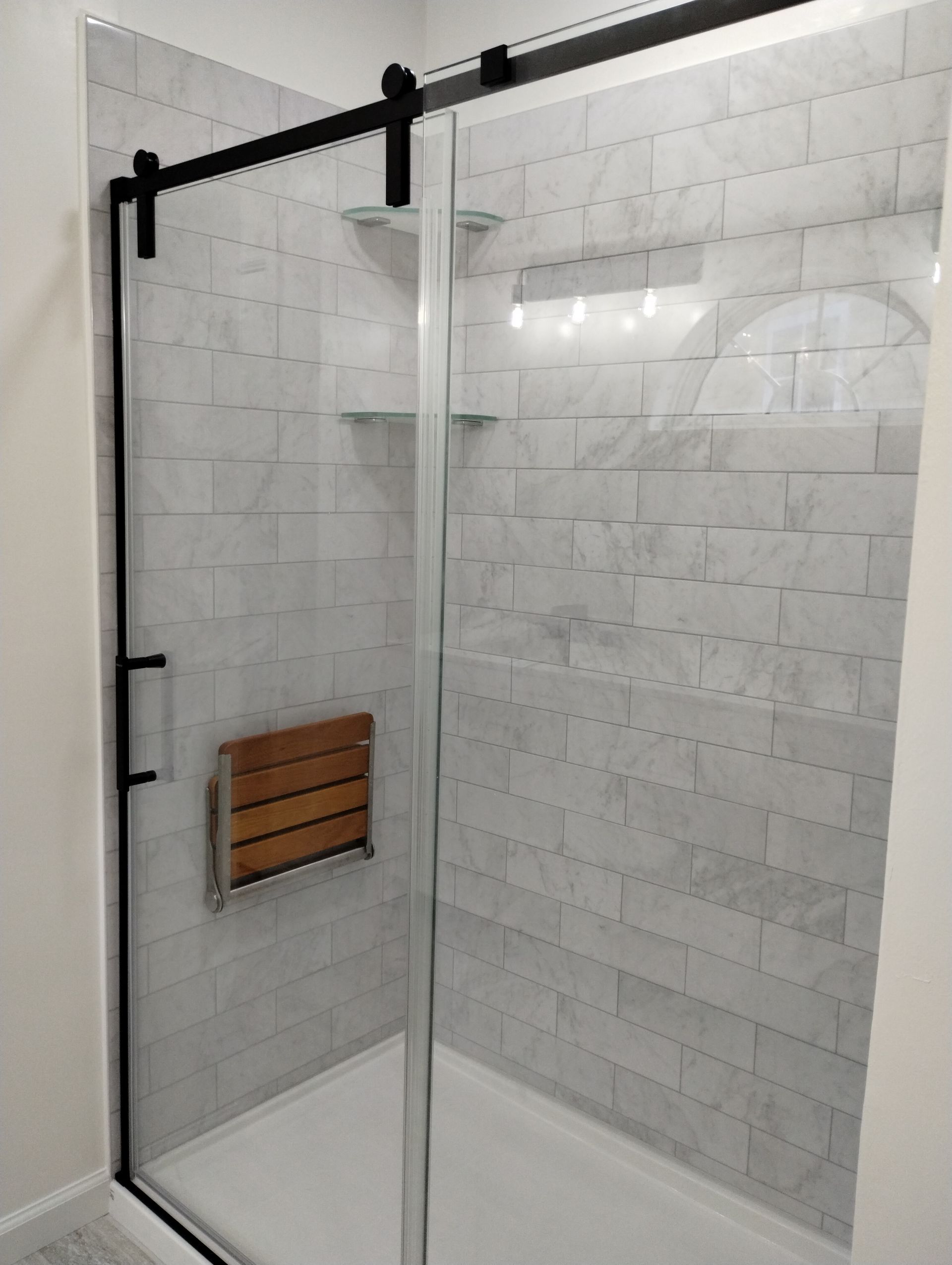 Glass shower enclosure with white tile walls and a wooden bench inside.