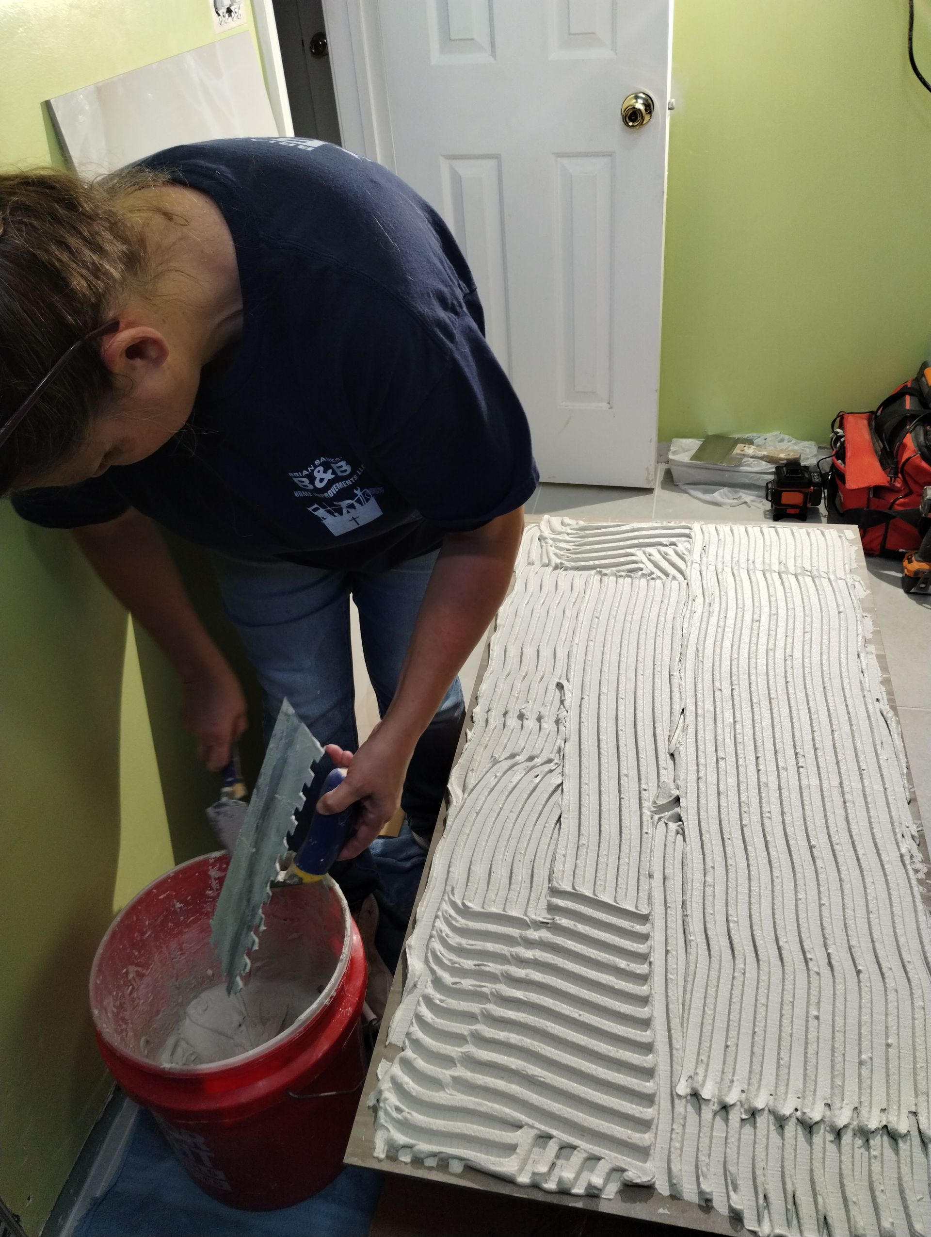 Person spreading white adhesive on a large board with a trowel in a room with a red bucket nearby