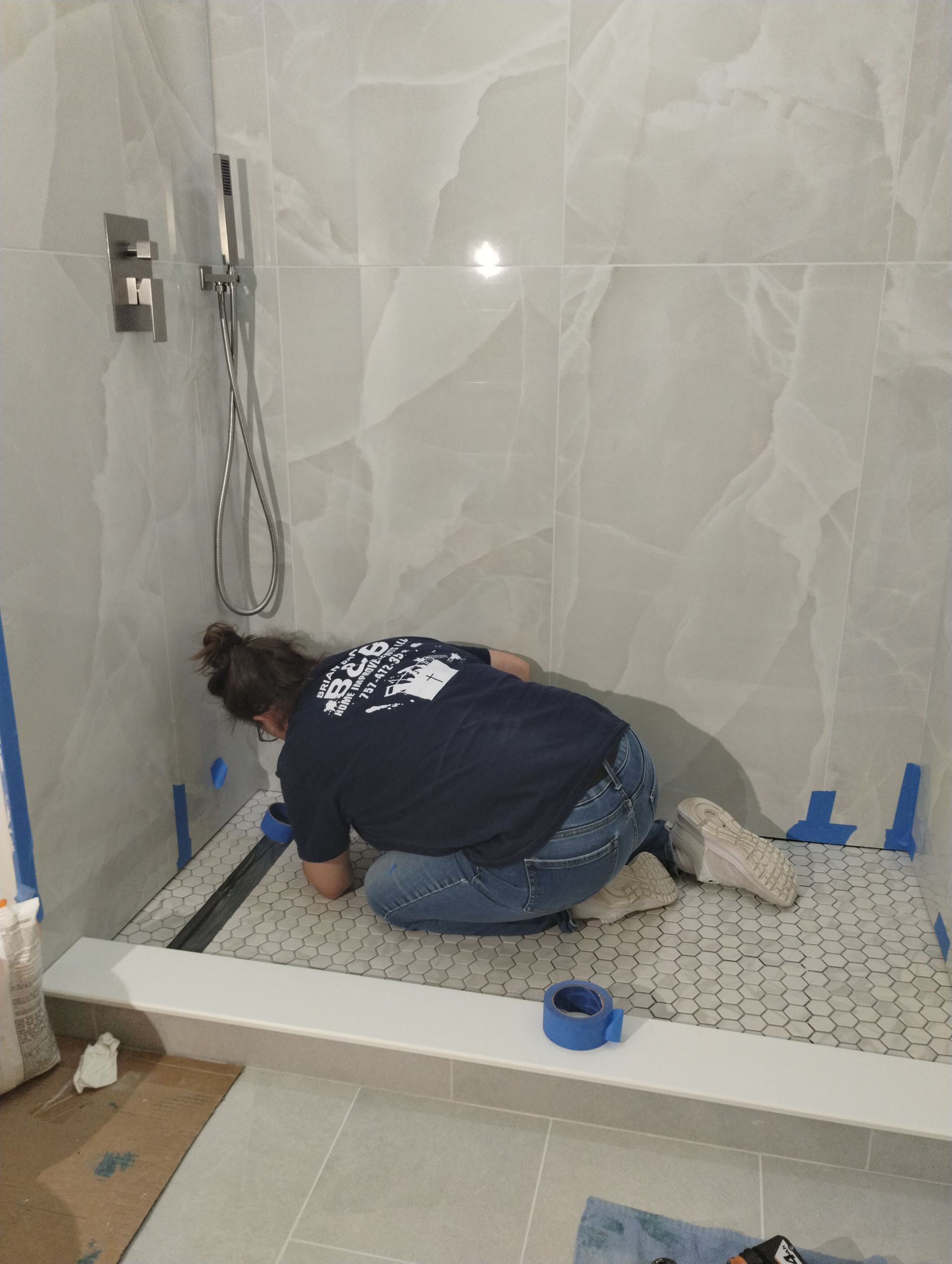 Worker installing mosaic tile in a shower stall with a handheld shower fixture and blue tape on the edges