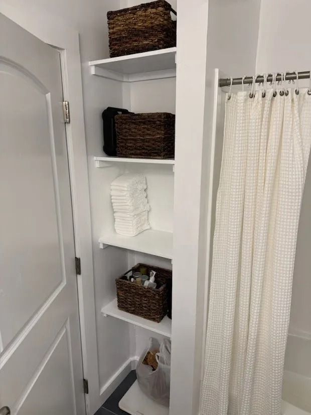 White bathroom shelf with wicker baskets, folded towels, and a shower curtain beside a frosted door