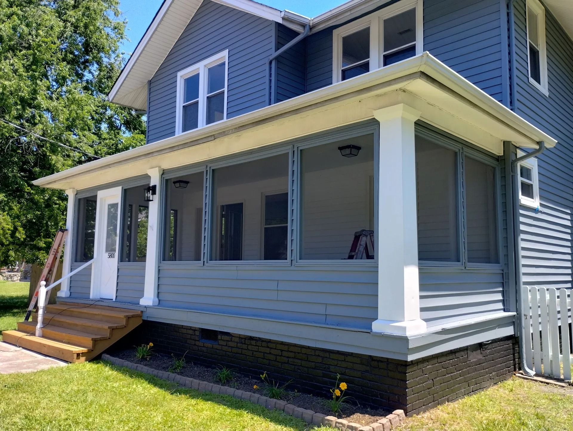 Blue two-story house with covered front porch, white trim, and wooden steps in a sunny yard