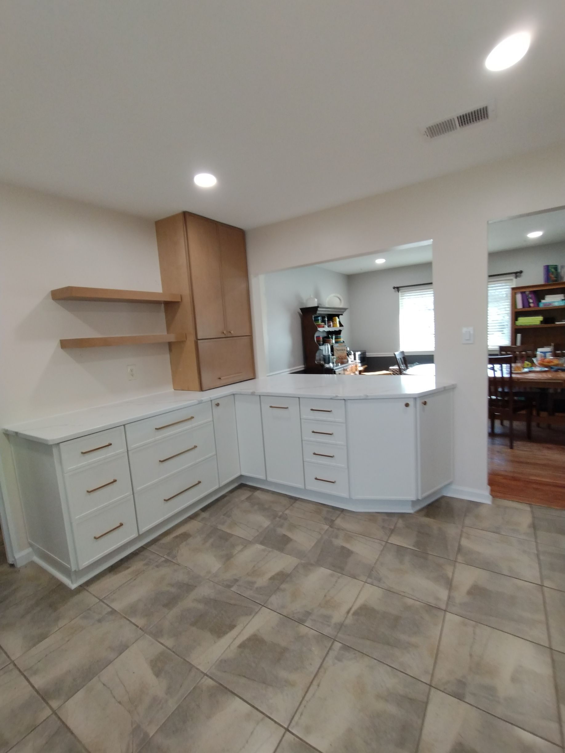 Bright kitchen with white cabinets, wood shelves, tiled floor, and a view into an adjoining dining area.