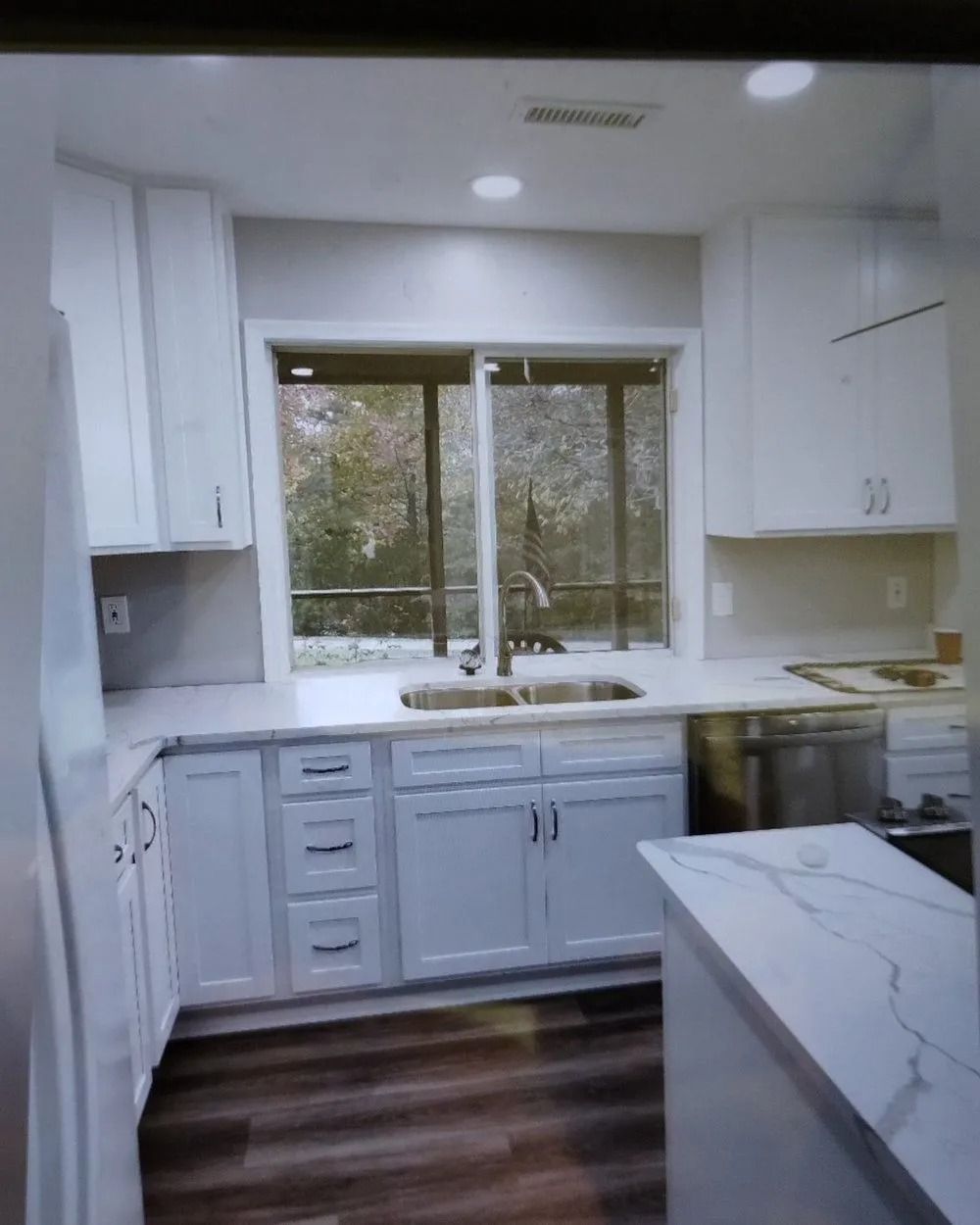 Modern white kitchen with window over sink, dark wood floor, and marble island countertop.