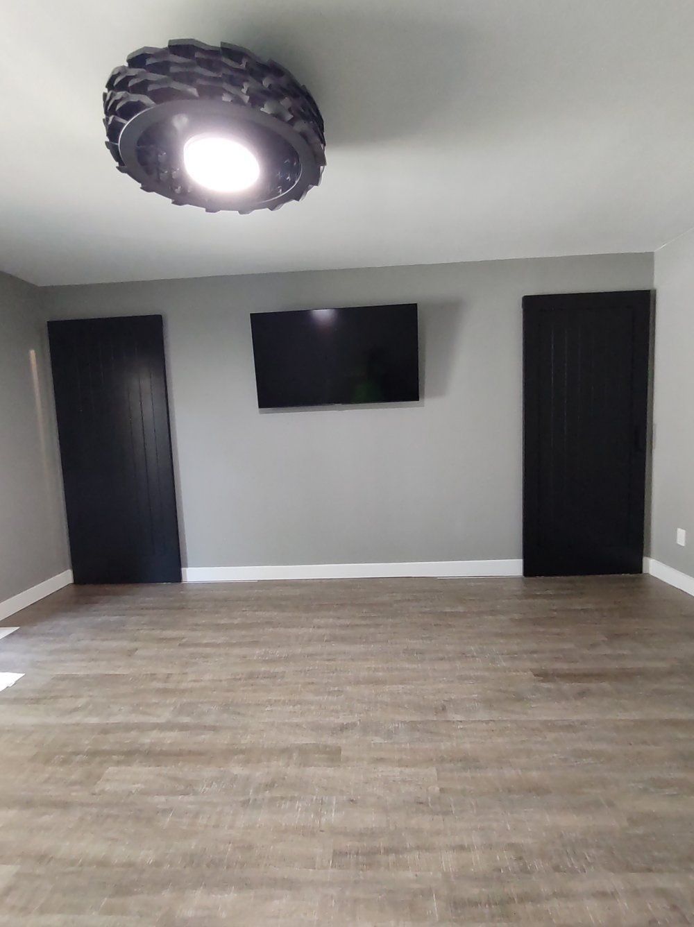 Empty gray room with wood flooring, two black doors, wall-mounted TV, and a ceiling light fixture.