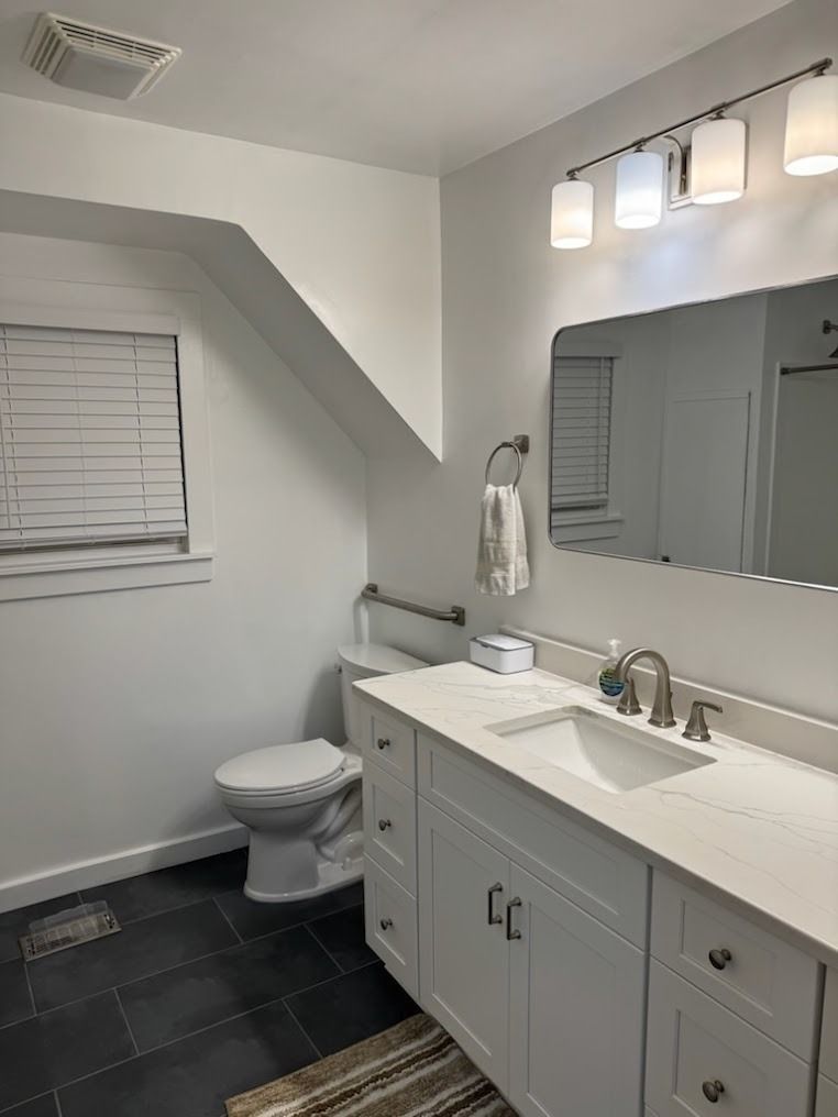 Modern bathroom with gray walls, white vanity, toilet, window, mirror, and dark tile floor