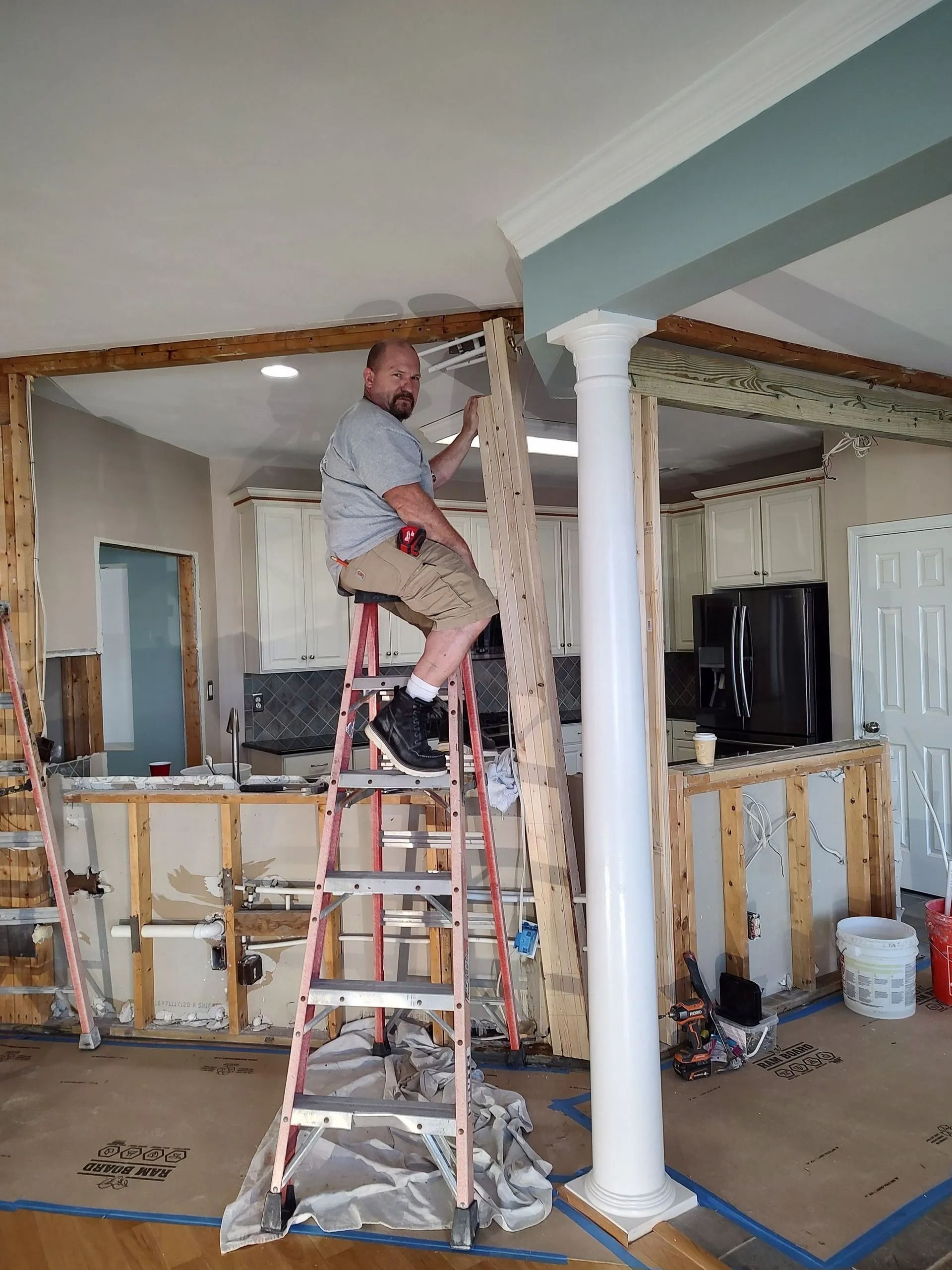 Worker on a ladder installing trim in a room under renovation, with exposed walls and columns.