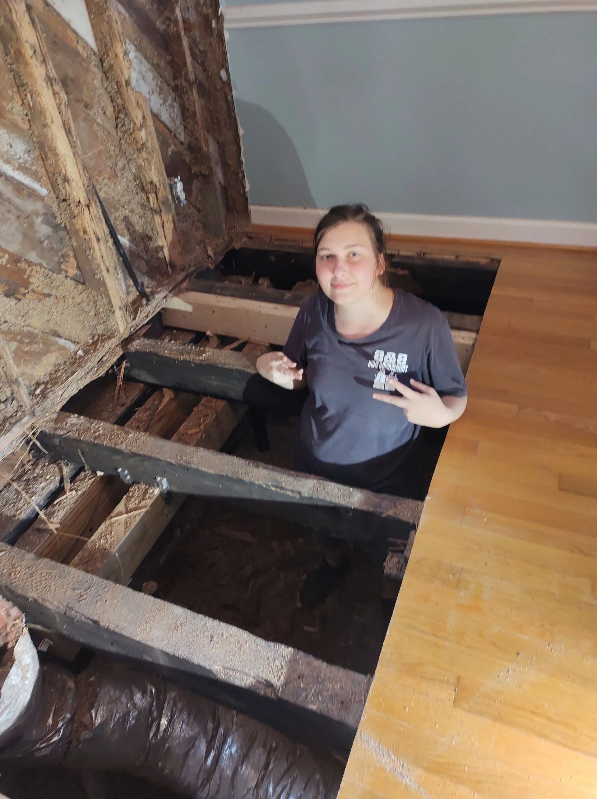 Person standing in an open floor joist area during home renovation, beside exposed beams and hardwood floor