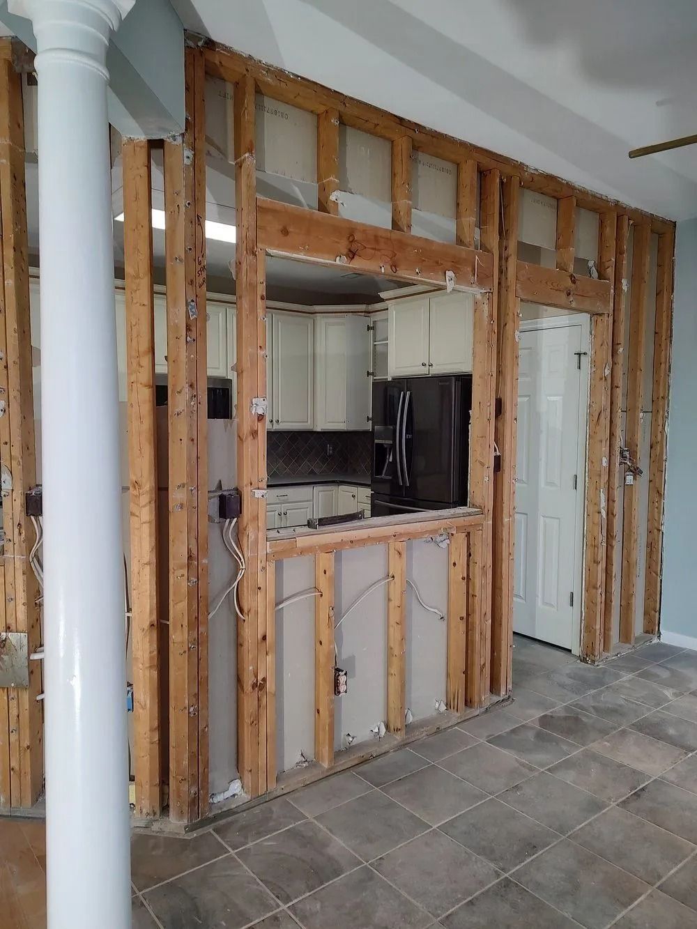 Unfinished kitchen wall with exposed studs and drywall around a serving opening and doorway