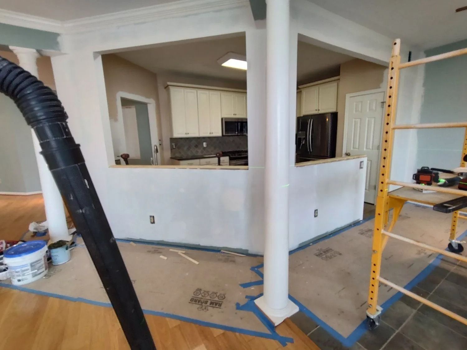 Renovated living room with white trim, open kitchen view, and a yellow ladder on a protected hardwood floor.