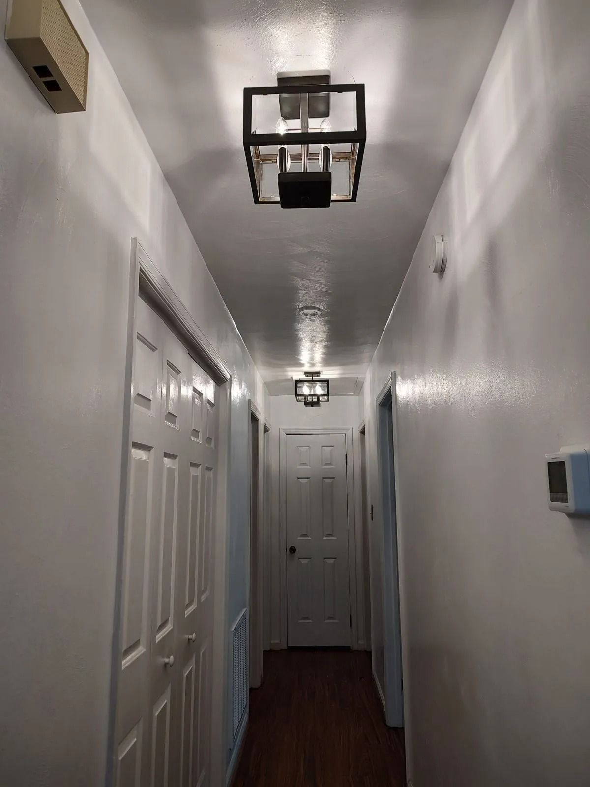 Narrow hallway with white walls, dark wood floor, ceiling lights, and closed doors along both sides.