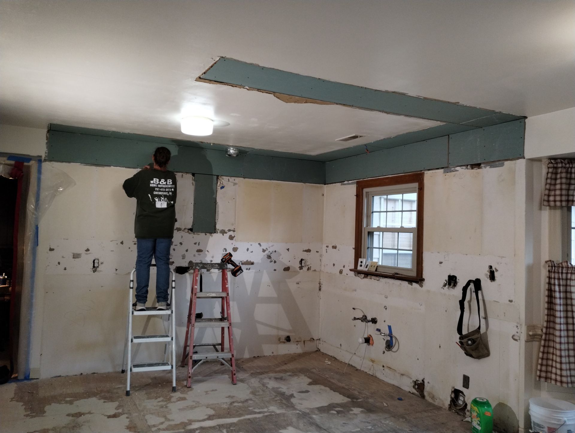 Person on a ladder painting a kitchen with white walls and teal trim during renovation