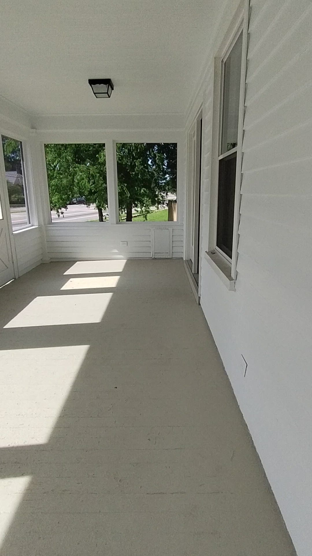 Empty screened-in porch with white walls, beige floor, and sunlight casting shadows through the windows.