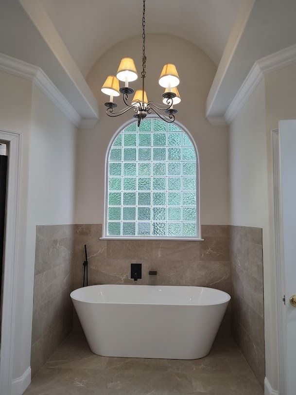 Elegant bathroom with freestanding white tub, arched glass block window, and chandelier lighting