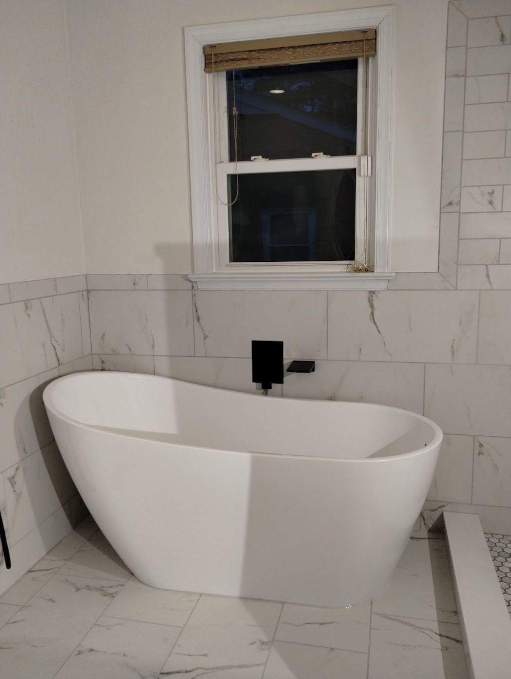 Modern white freestanding bathtub in a tiled bathroom beneath a small window