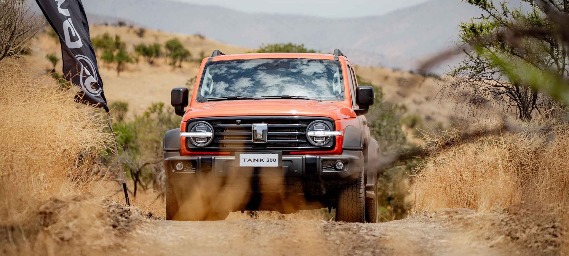 Camioneta todoterreno naranja circulando por un terreno desértico polvoriento con montañas al fondo.