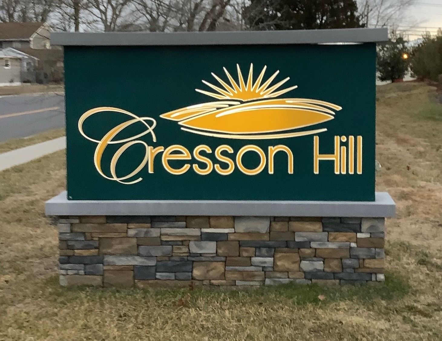 Cresson Hill sign, green background with gold text and sunrise graphic, on stone base.