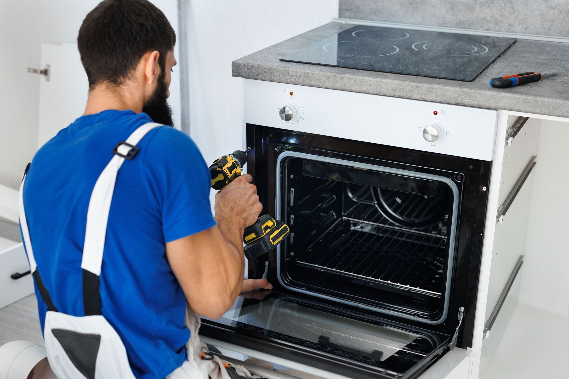 Technician in blue shirt installing an oven in a kitchen, holding a power drill.