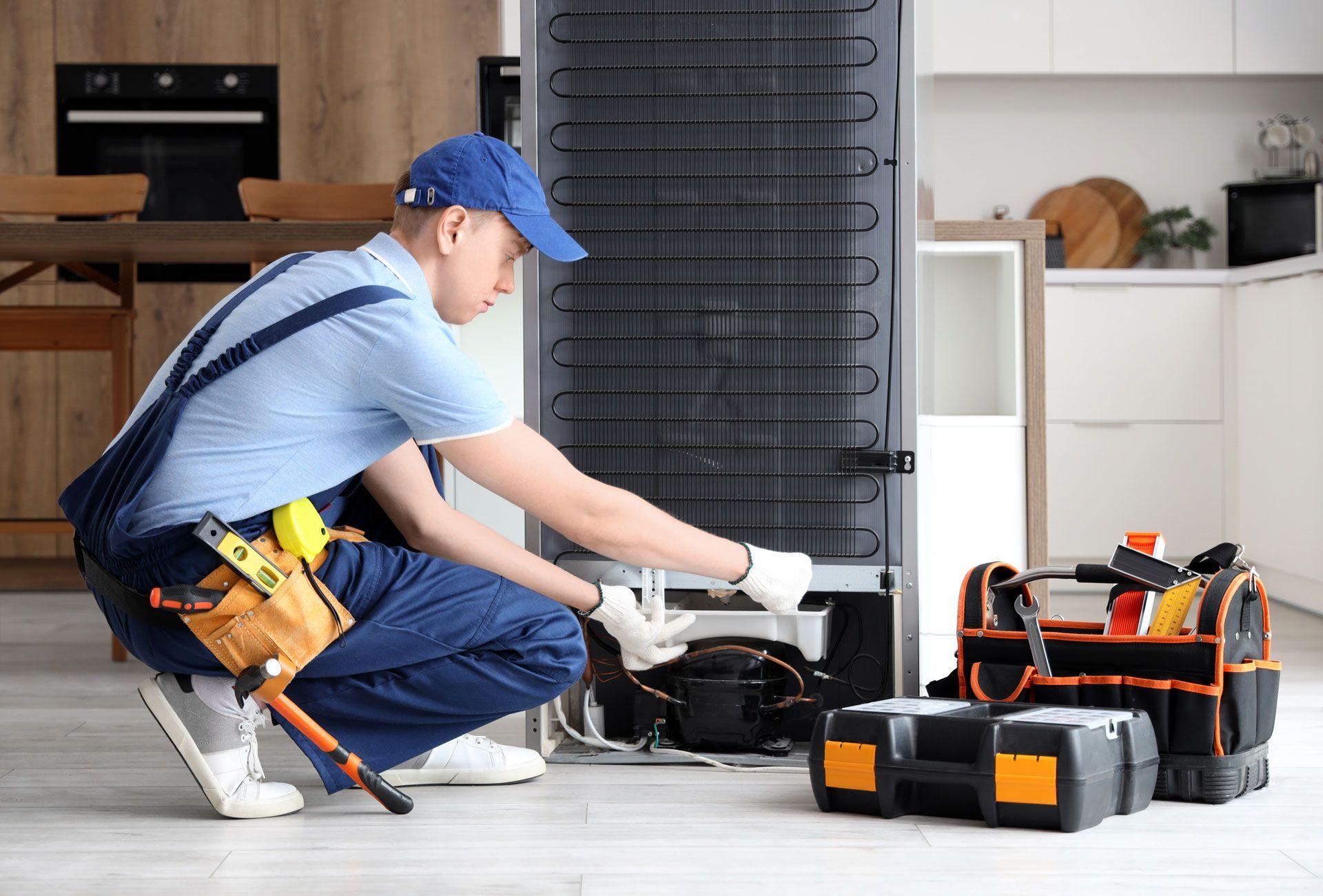 Technician kneels beside a refrigerator, repairing it with tools in a kitchen.