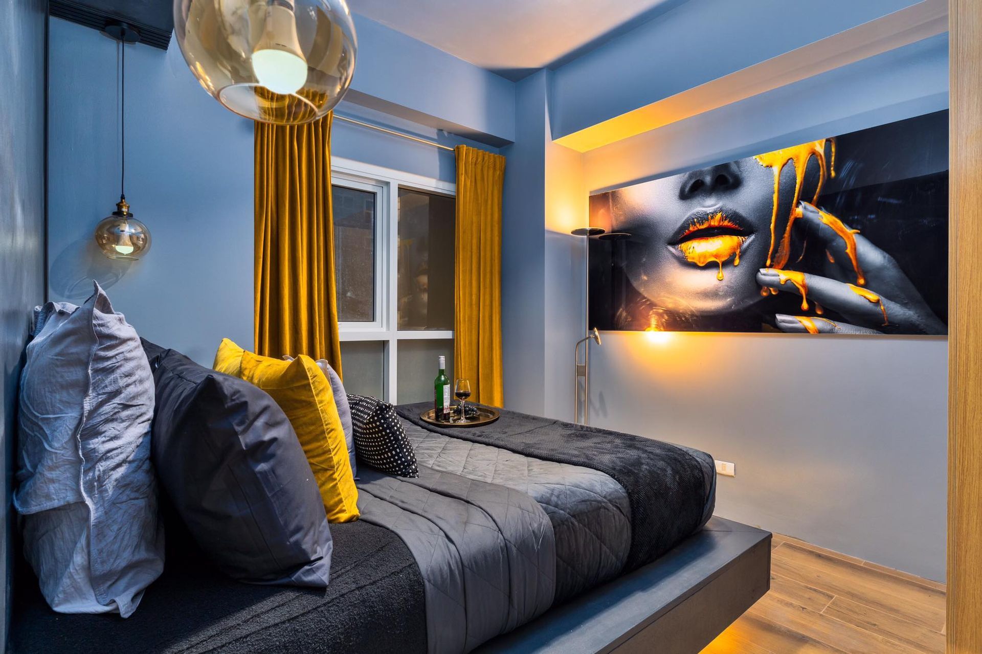 A bedroom with a large bed in grays and oranges and a backlit painting on the wall. The Den at Bayshore Luxe by Prime, Manila