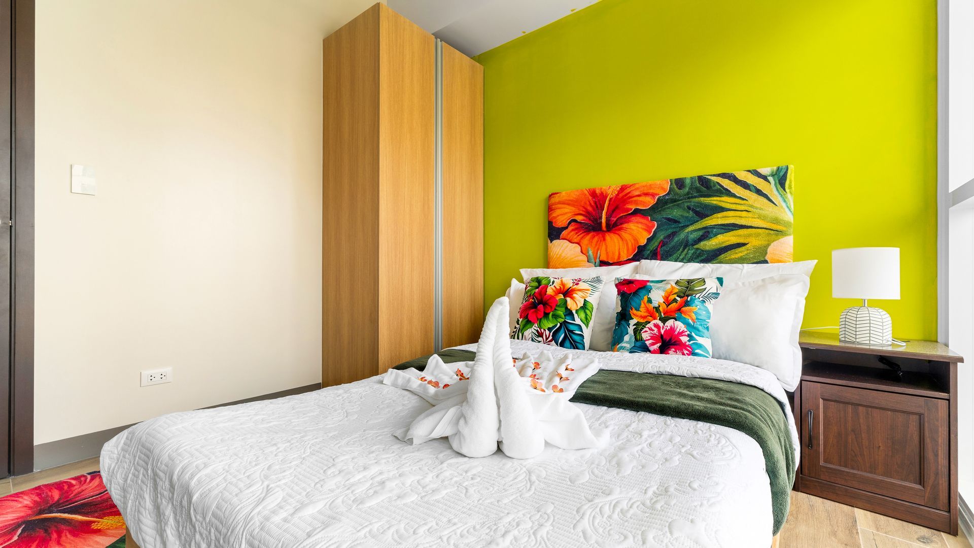 Bed with white bed linens and tropical cushions, against a bright colour wall