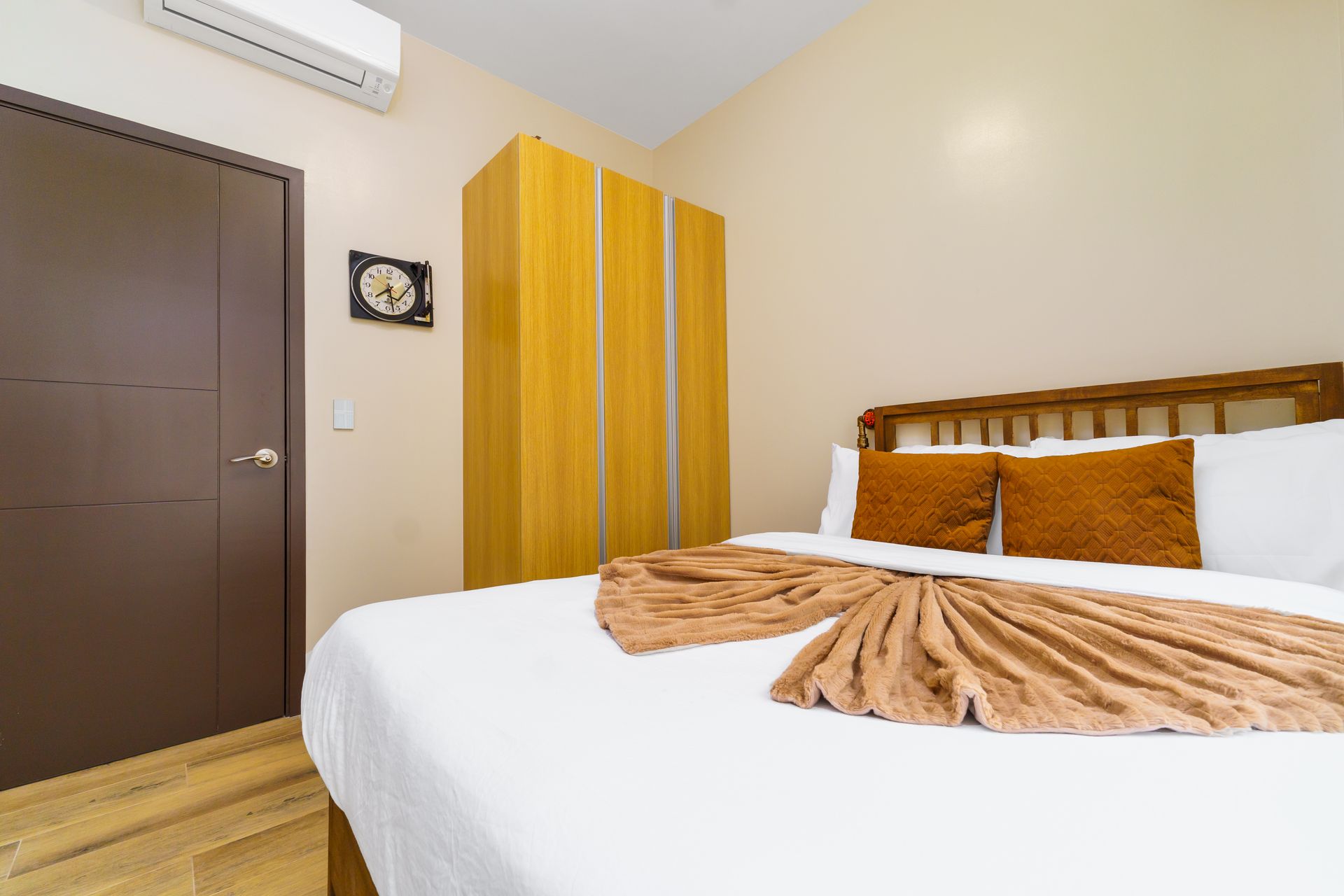 Wooden Bed frame, bed with white pillows and linens, and rust brown cushions, wooden wardrobe in the background