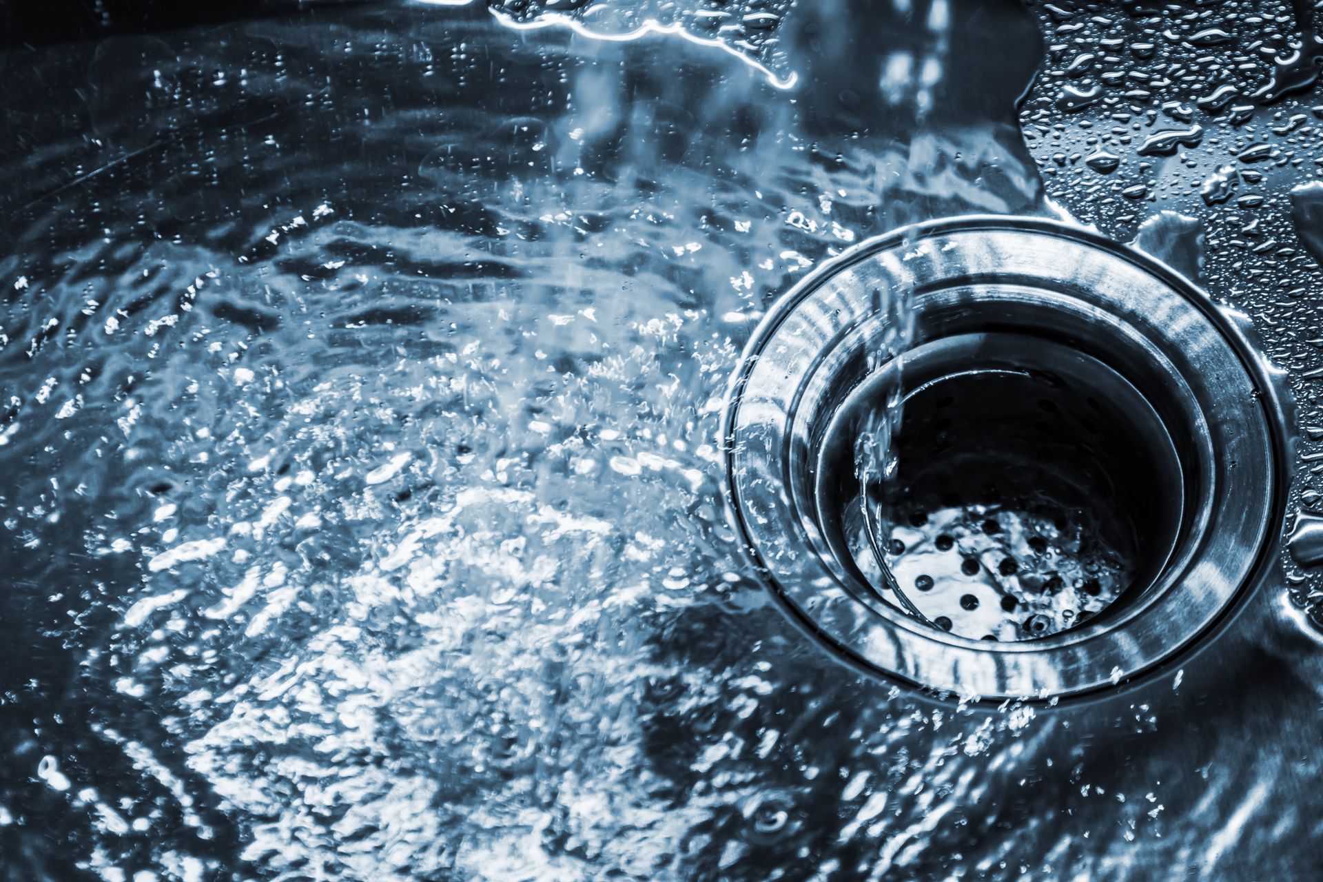 Water swirling into a stainless steel sink drain with splashing droplets.