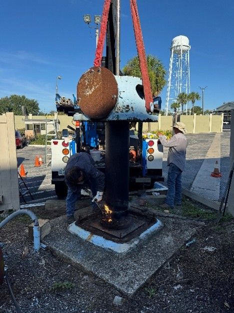 Photograph showing two workers performing maintenance on a large industrial valve or pipe outdoors, with equipment and vehicles in the background.