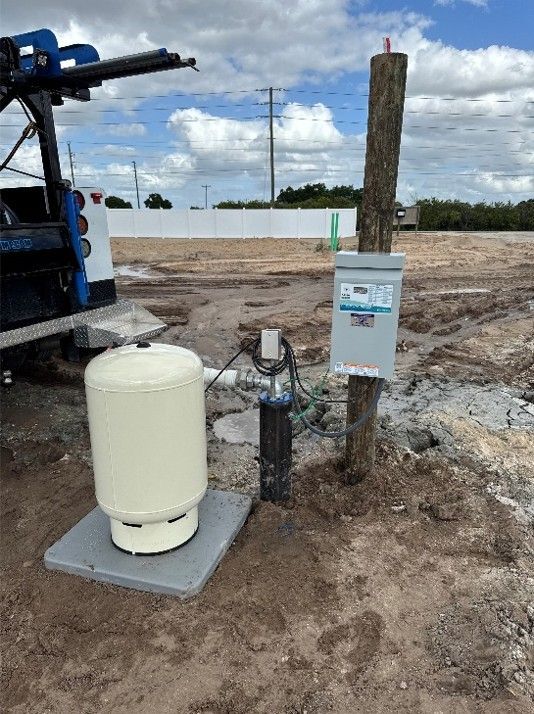 Photograph of an outdoor propane tank setup at a construction or utility site, showing a white cylindrical propane tank mounted on a concrete slab next to a wooden post with an attached control box and connected piping.