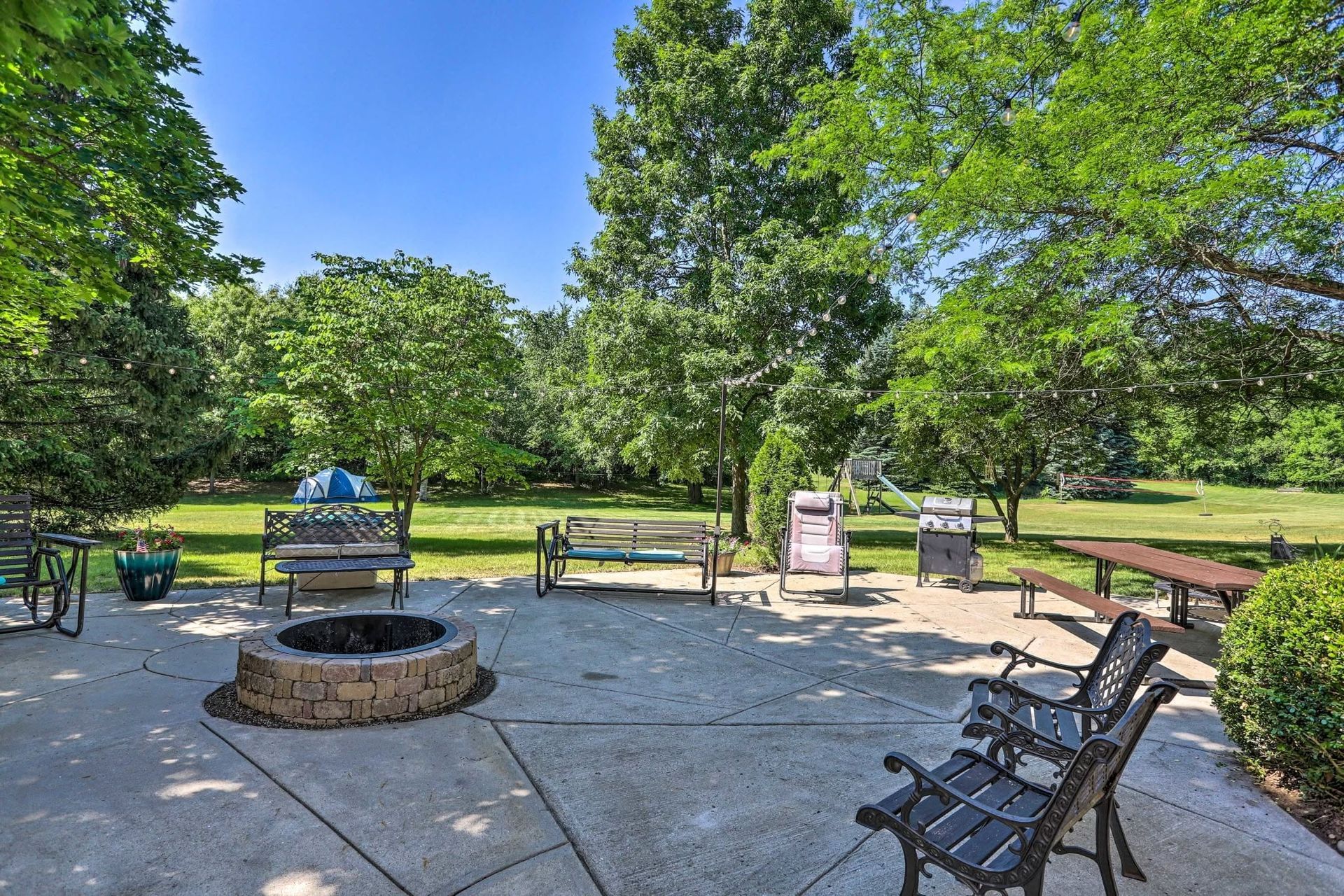 Park patio with fire pit, benches, and chairs surrounded by green trees and lawn.