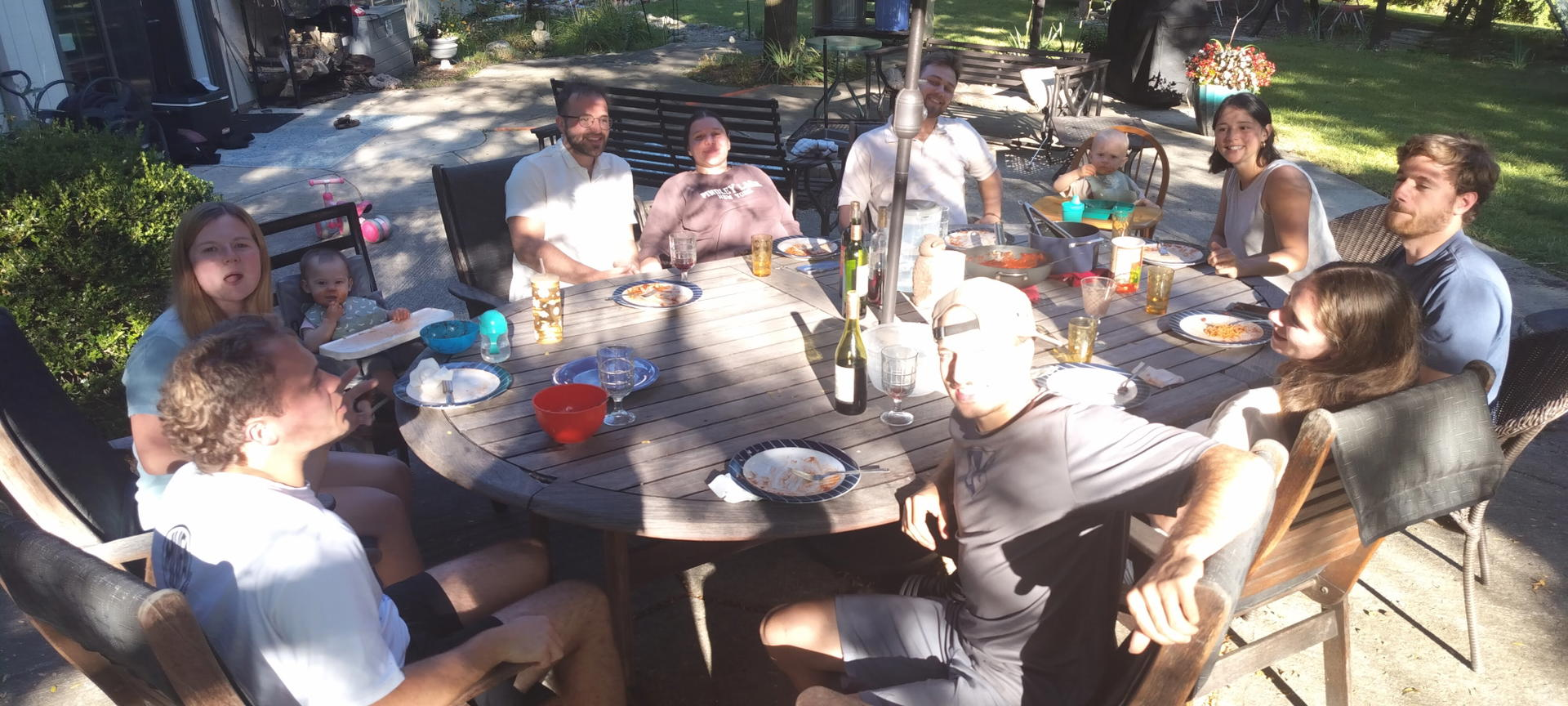 People gathered around a round outdoor table in a sunny backyard, eating and chatting.