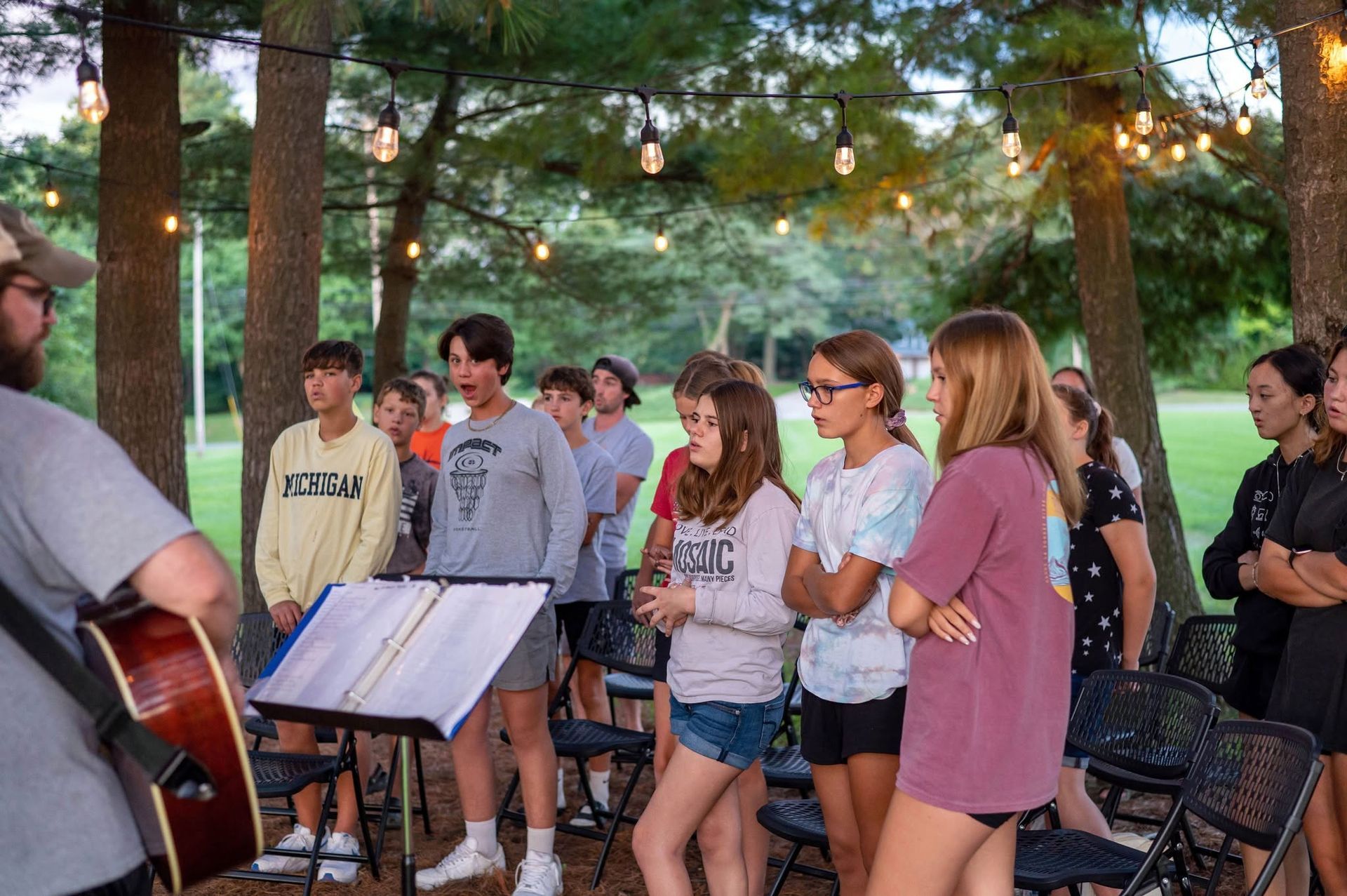 Group of people singing outdoors at dusk, with a guitarist and music stand among trees and string lights.