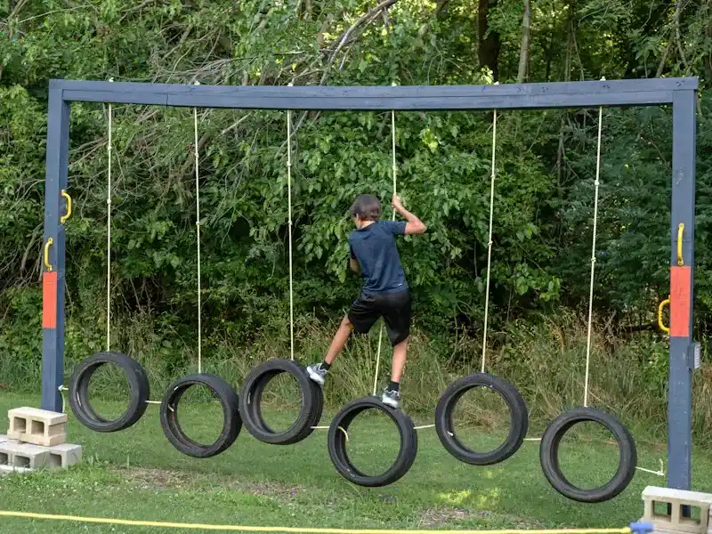 Person crossing hanging tires in an outdoor obstacle course, wearing dark clothes and a cap.