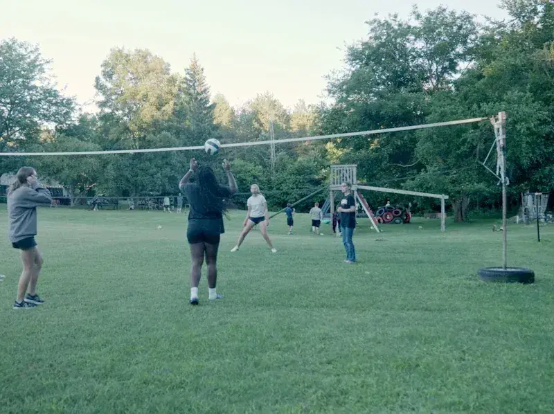 People playing volleyball on a grassy field at dusk, with a net and trees in the background