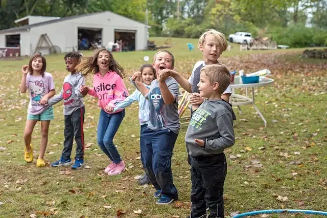 Children standing and smiling outdoors near a house, with water splashing and leaves on the ground.