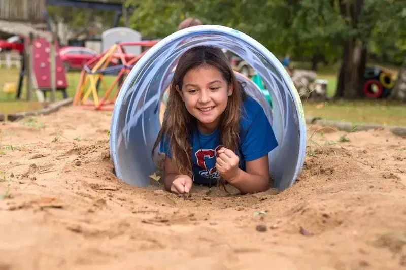 Child smiling inside a blue tunnel tube at a playground, lying on sandy ground.