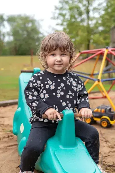 Child sitting on a green playground toy, wearing a black floral shirt, outdoors at a park