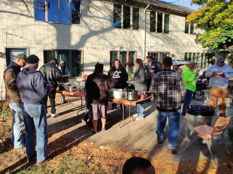 People gathered outdoors around tables and serving pots in a sunny courtyard by a building.