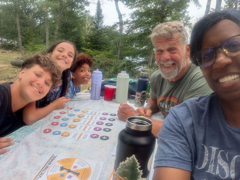 Family gathered around a picnic table outdoors, smiling with drinks and a board game.